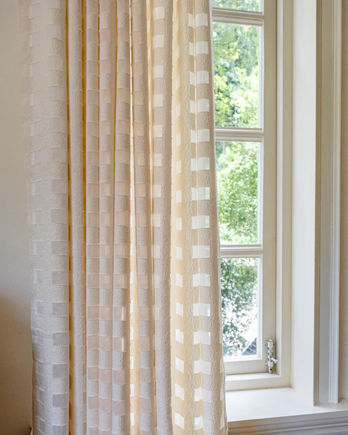 A full-length view of the Aria sheer check curtain hanging from a black rod next to a window.