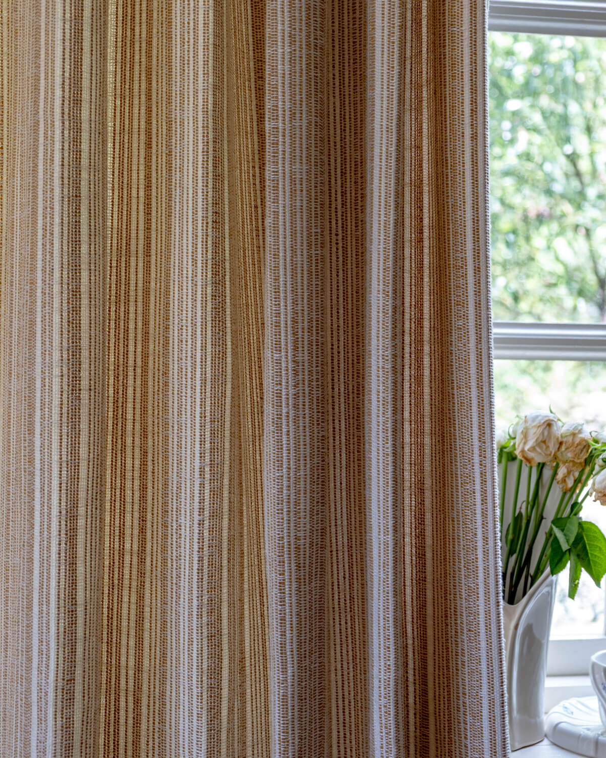 The light-filtering fabric of the Thea striped curtain hanging in front of a window with flowers.