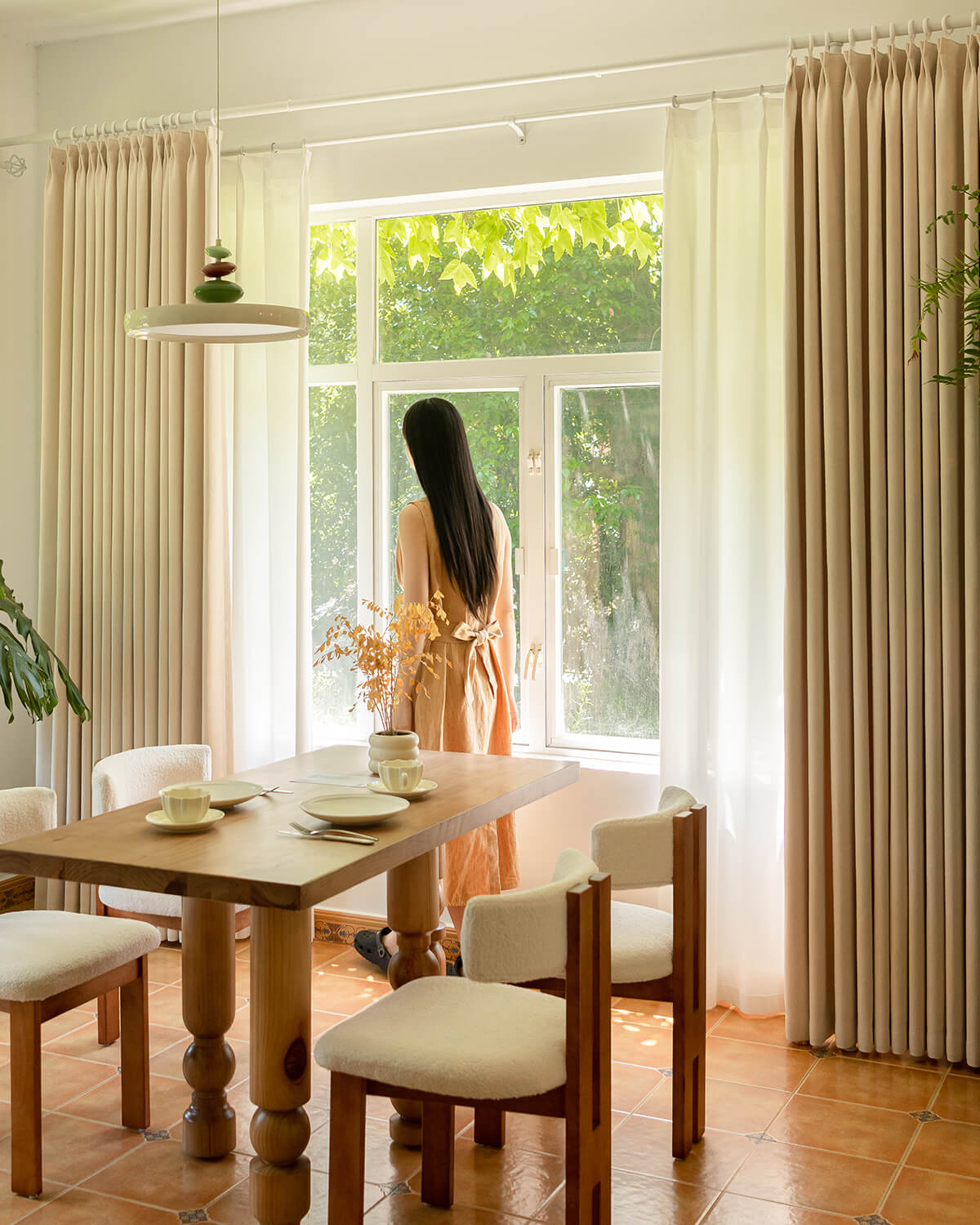 A woman looking out a window framed by Mell beige Chenille blackout curtains in a dining room.