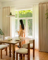 A woman looking out a window framed by Mell beige Chenille blackout curtains in a dining room.