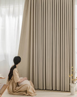 A woman sitting on the floor next to the floor-length Fuzz beige Chenille blackout drapes.
