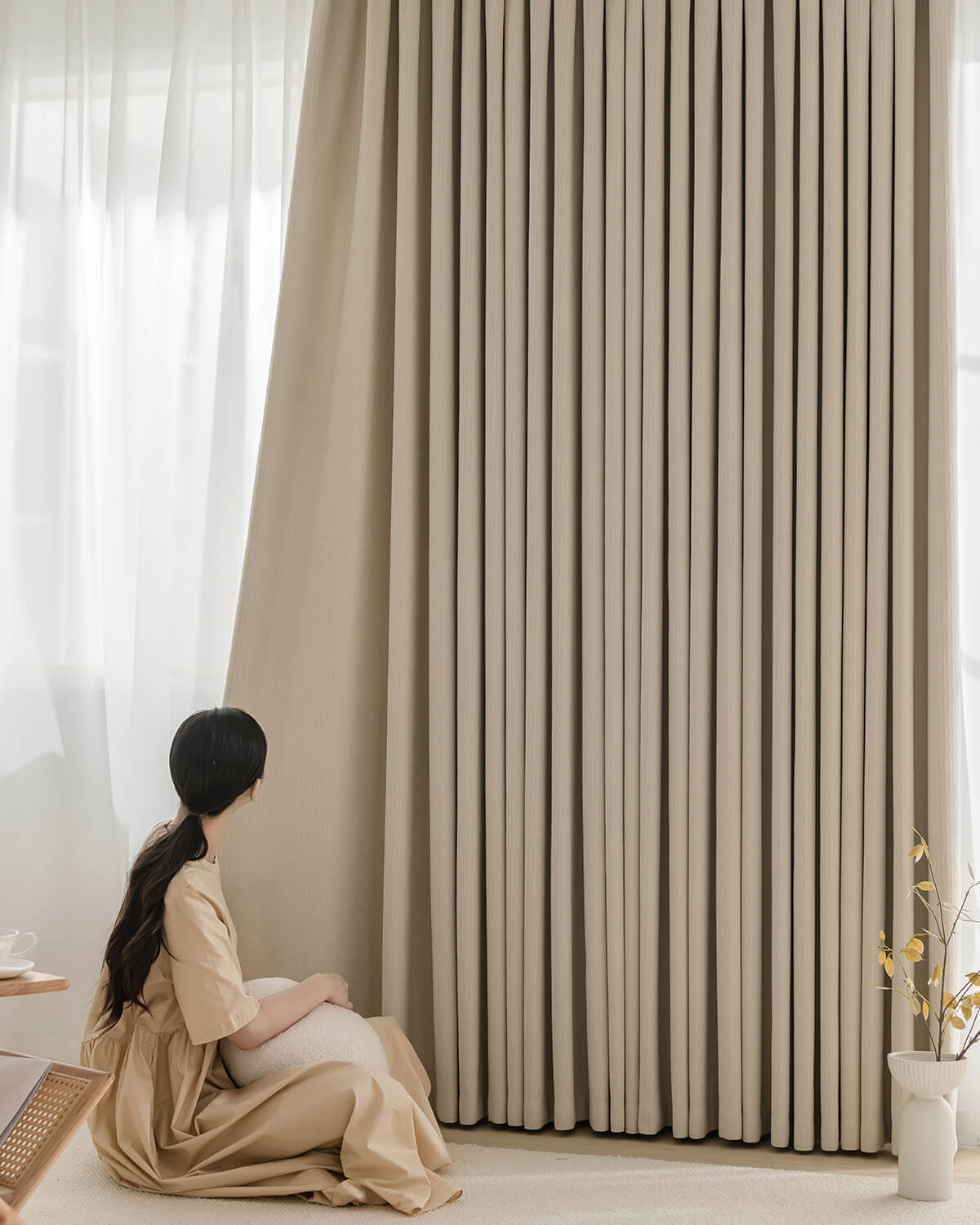 A woman sitting on the floor next to the floor-length Fuzz beige Chenille blackout drapes.