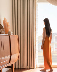 Woman in orange dress standing by floor-length beige waffle weave curtains with ruffle trim in a sunlit room.