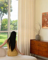 A woman in a yellow dress looking out a window dressed with Tina thick linen blend light-filtering curtains.