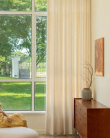 Tina pinch pleated linen blend drapes framing a large window with a view of green trees in a sunlit room.